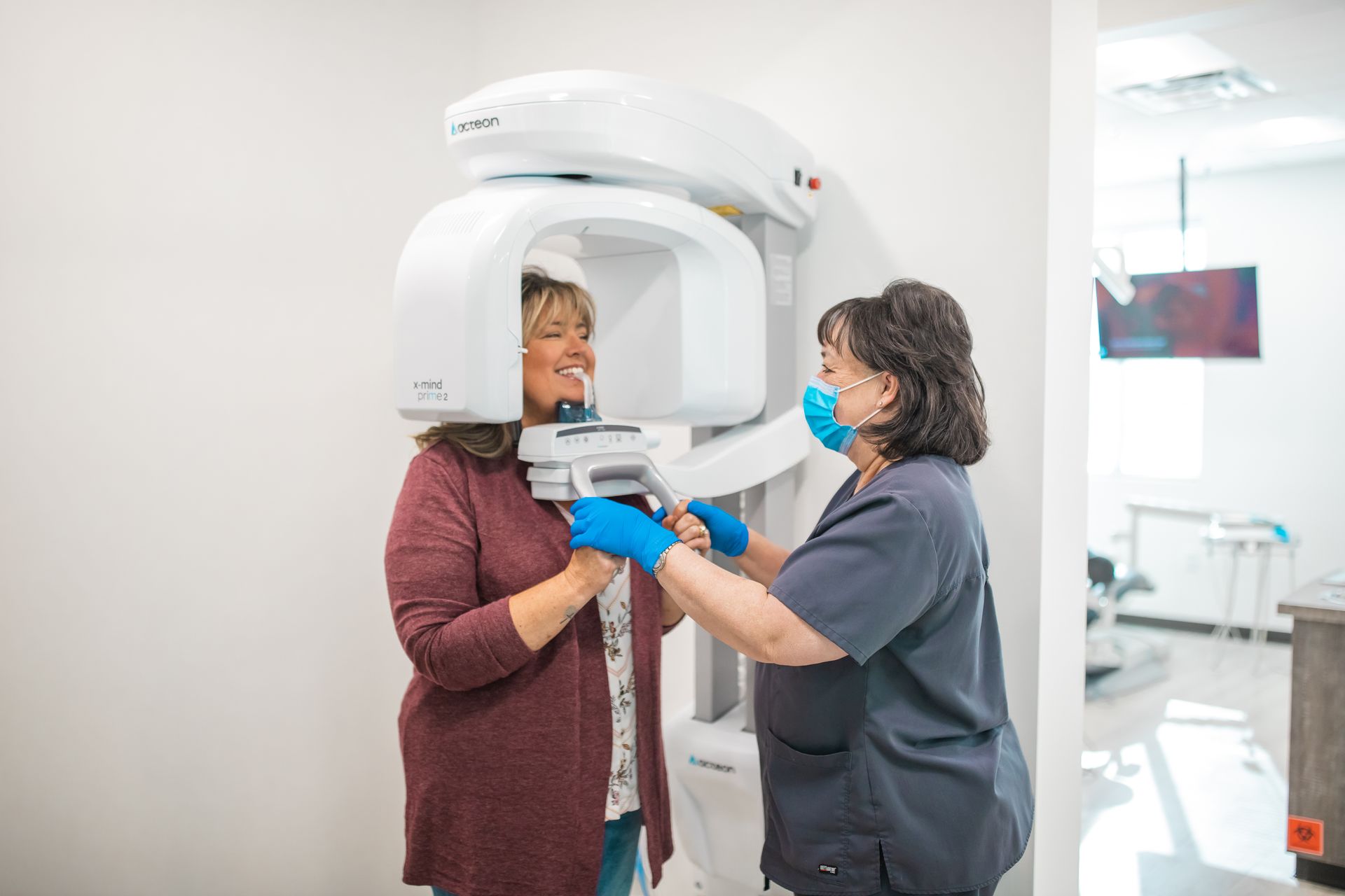 Woman getting dental X-ray. Dental assistant adjusts machine, both wearing masks. Bright room.