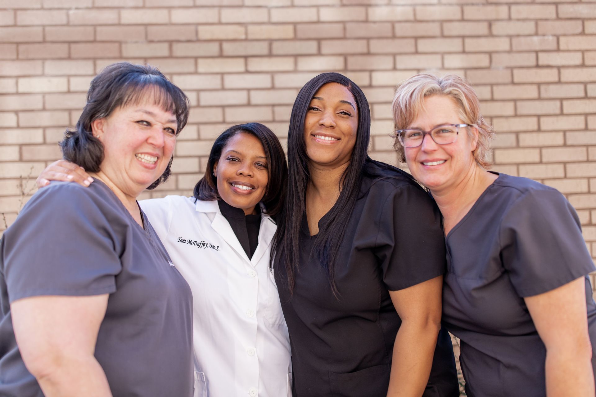 Four people in medical scrubs and lab coat smile together in front of a brick wall.