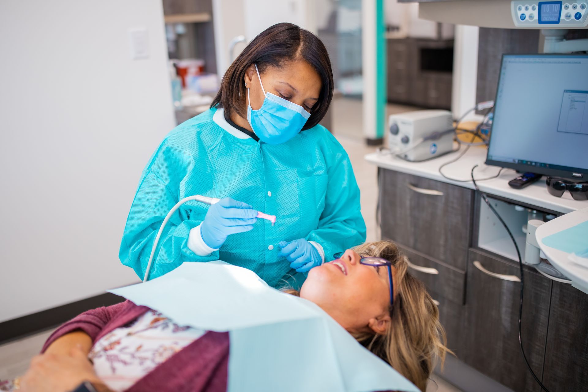 Dentist in blue scrubs and mask examining a patient's teeth in a dental office.