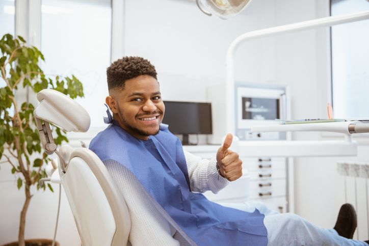 Man in dental chair giving thumbs up, smiling. Blue bib, bright dental office.