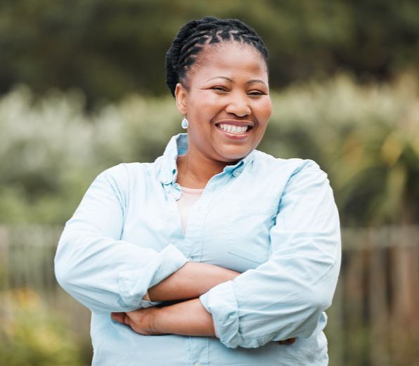 Woman with arms crossed, smiling. Wearing a light blue button-down shirt outdoors.