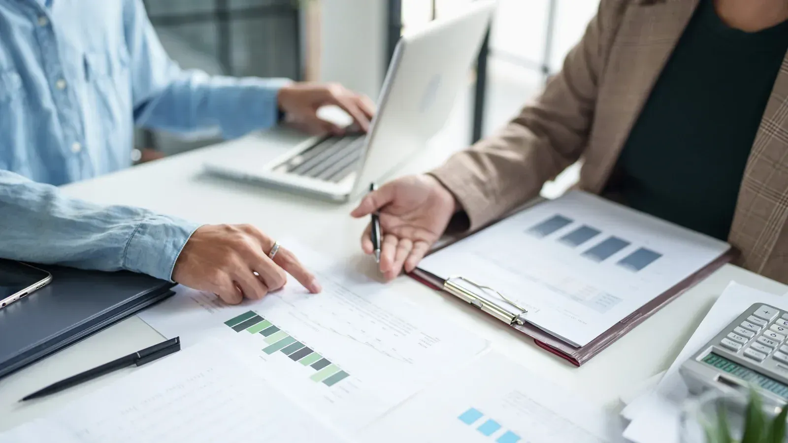 Two people reviewing financial documents, one using a laptop, on a white desk.