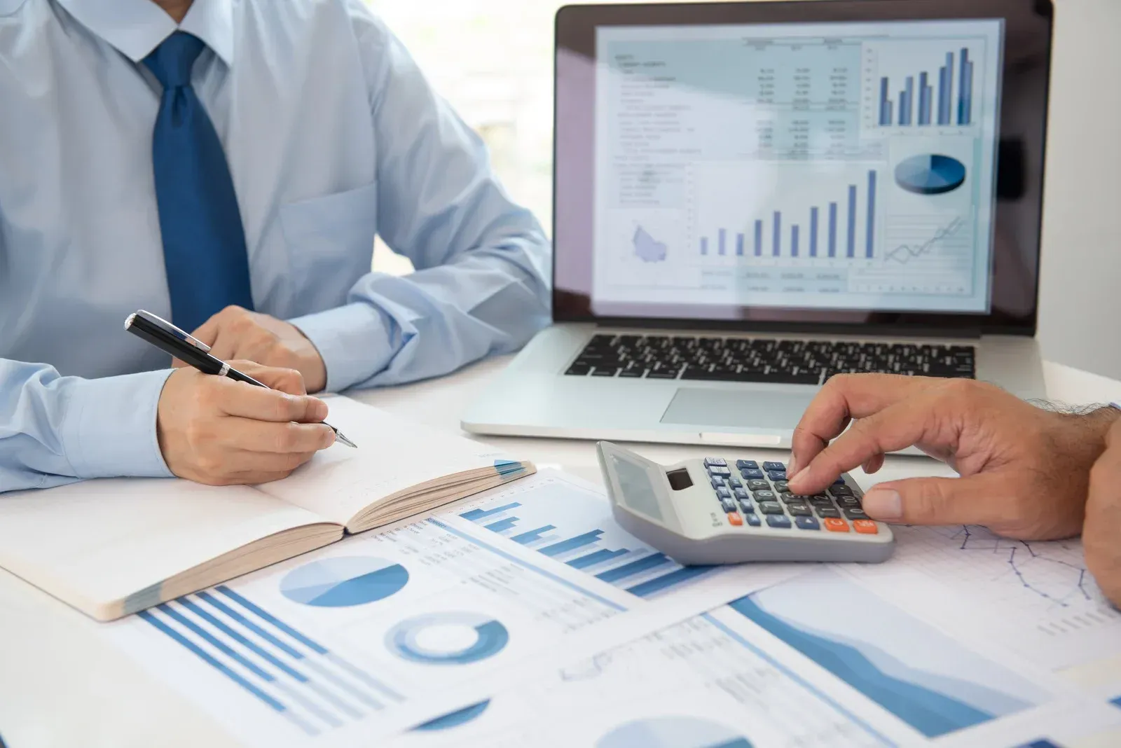 Two people at a desk review financial data on a laptop, notebook, calculator, and printed charts.