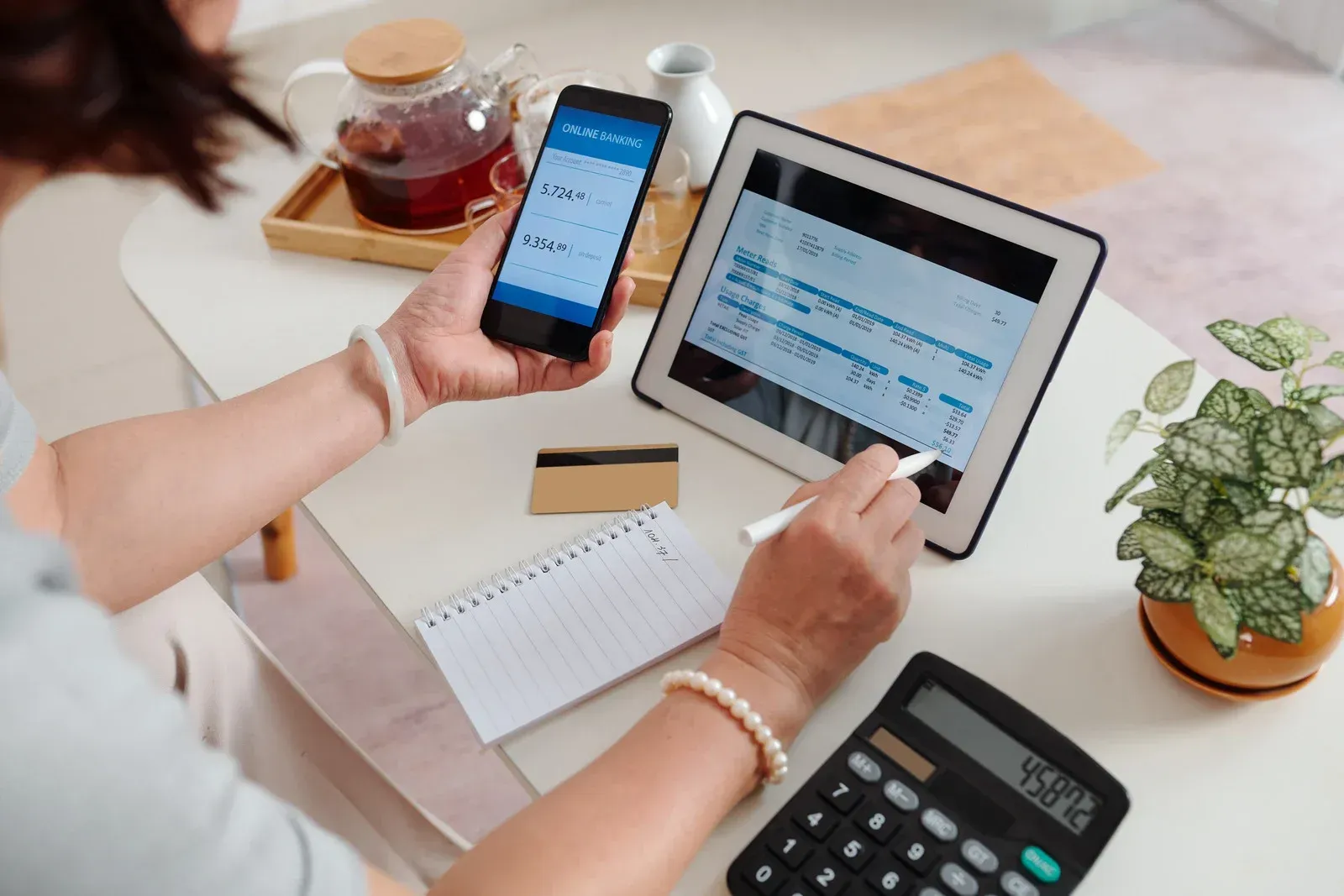 Woman using a tablet and phone, with a calculator, notepad, and credit card on a table.