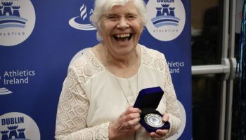An elderly woman is holding a medal and smiling.