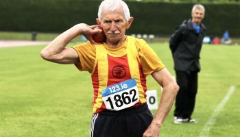 An older man wearing a yellow and red shirt with the number 1862 on his chest is throwing a ball.