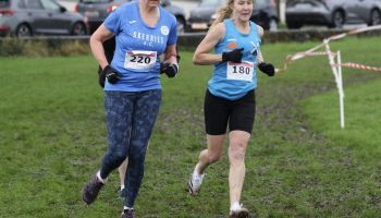 Two women are running in a cross country race on a grassy field.