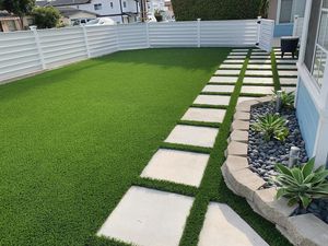 Artificial grass backyard with a stone walkway, white fence, and small succulents.