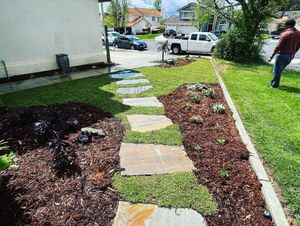 Stone path through a landscaped yard with mulch, grass, and a person watering the lawn.
