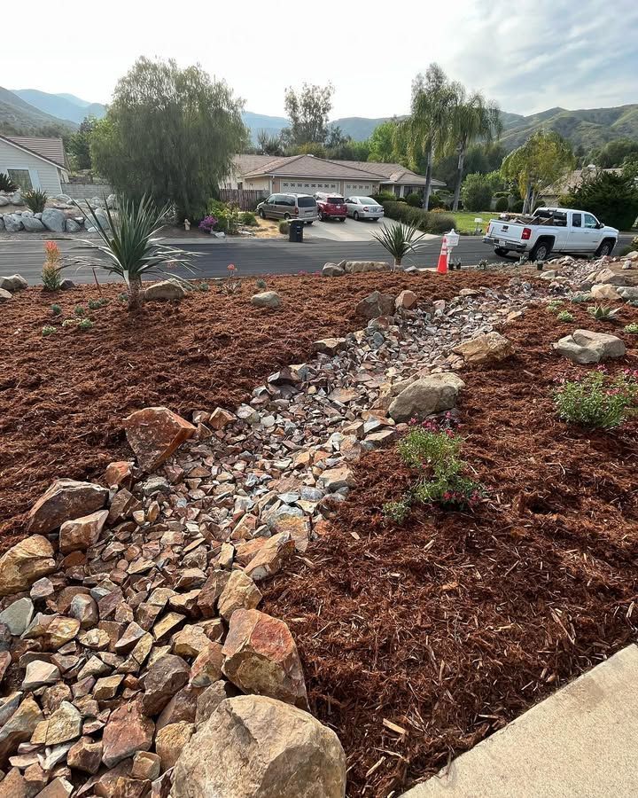 A front yard with a dry creek bed and brown mulch, houses in the background.