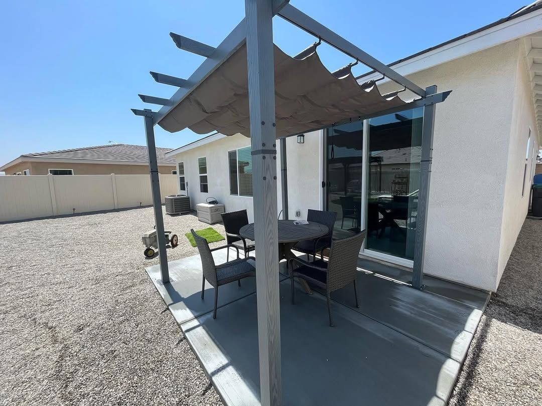 Patio with table and chairs under a pergola, next to a house with a sliding glass door.