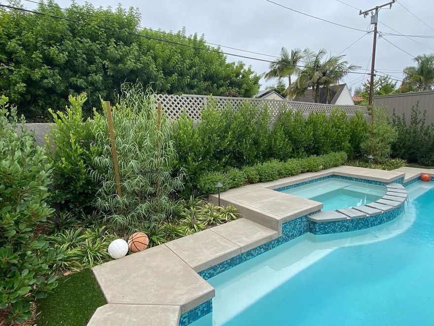 Pool with blue tiles, hot tub, concrete steps, and lush green landscaping.