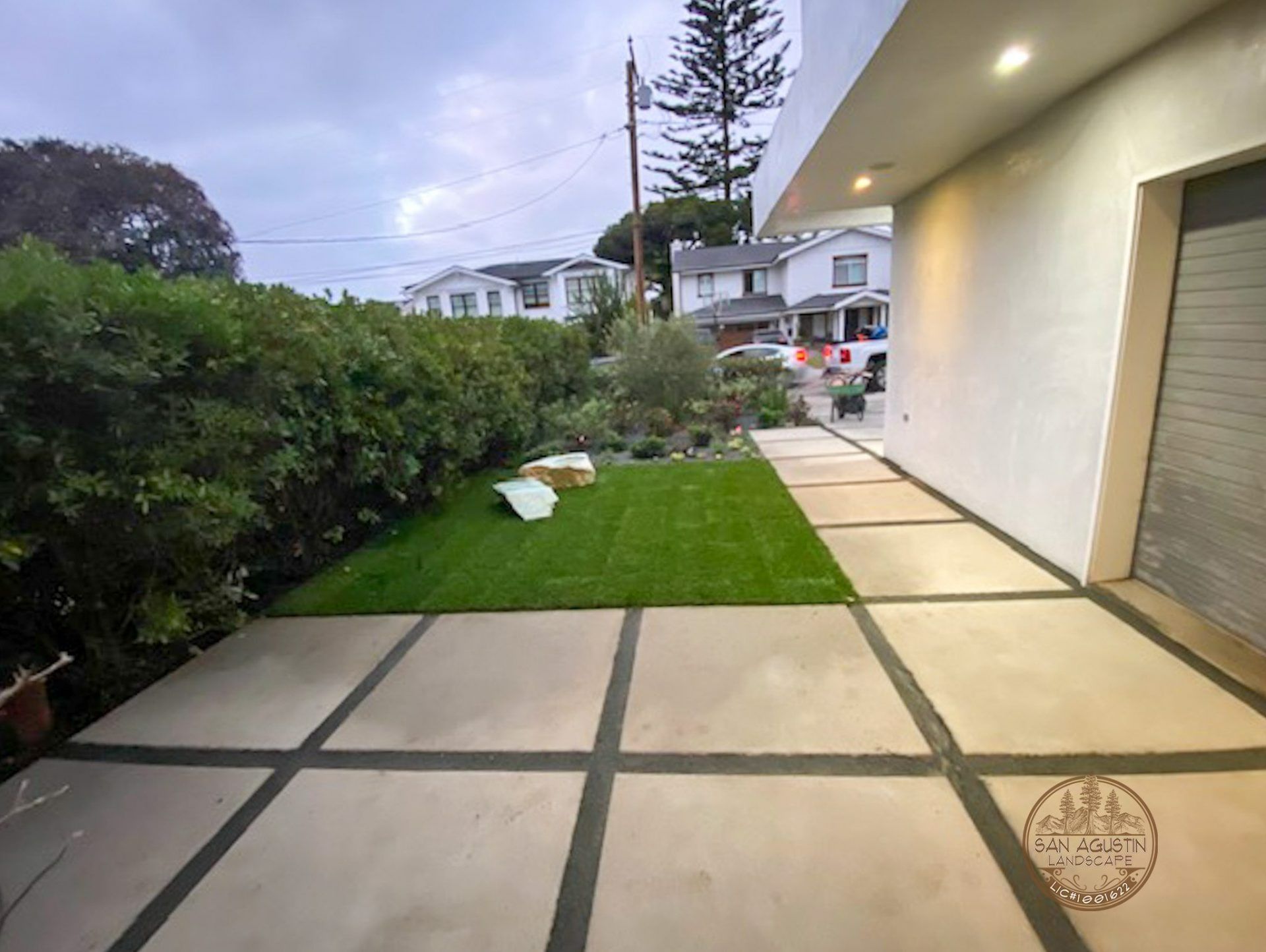 Patio with concrete pavers, green grass, and garden, near a white modern house. Cloudy sky.