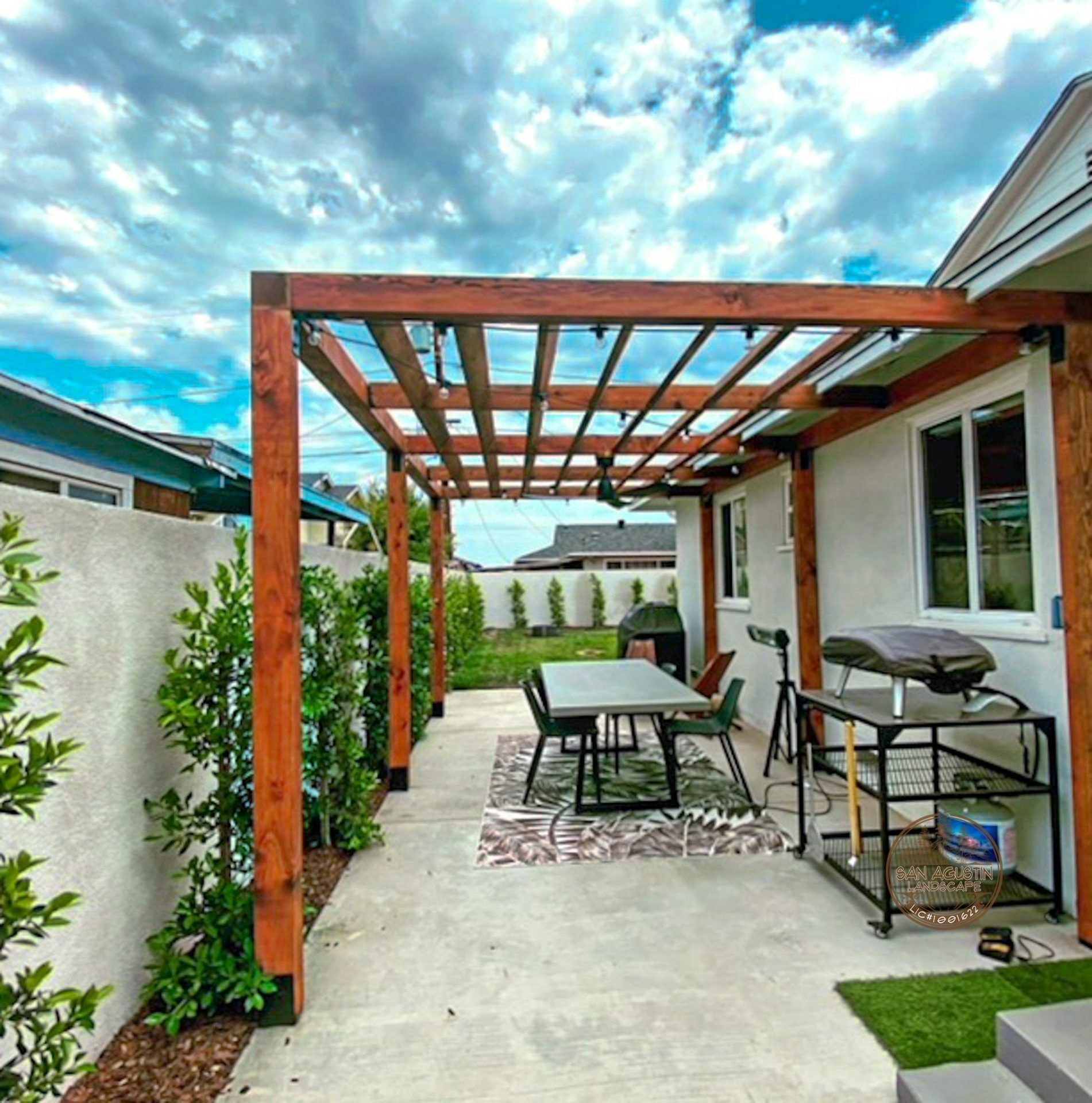 Wooden pergola over a patio with a table, chairs, and grill; a partially cloudy sky is above.