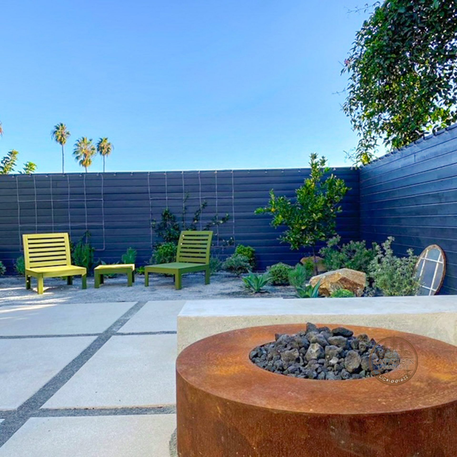 Outdoor patio with fire pit, concrete pavers, green chairs, and a dark fence under a blue sky.