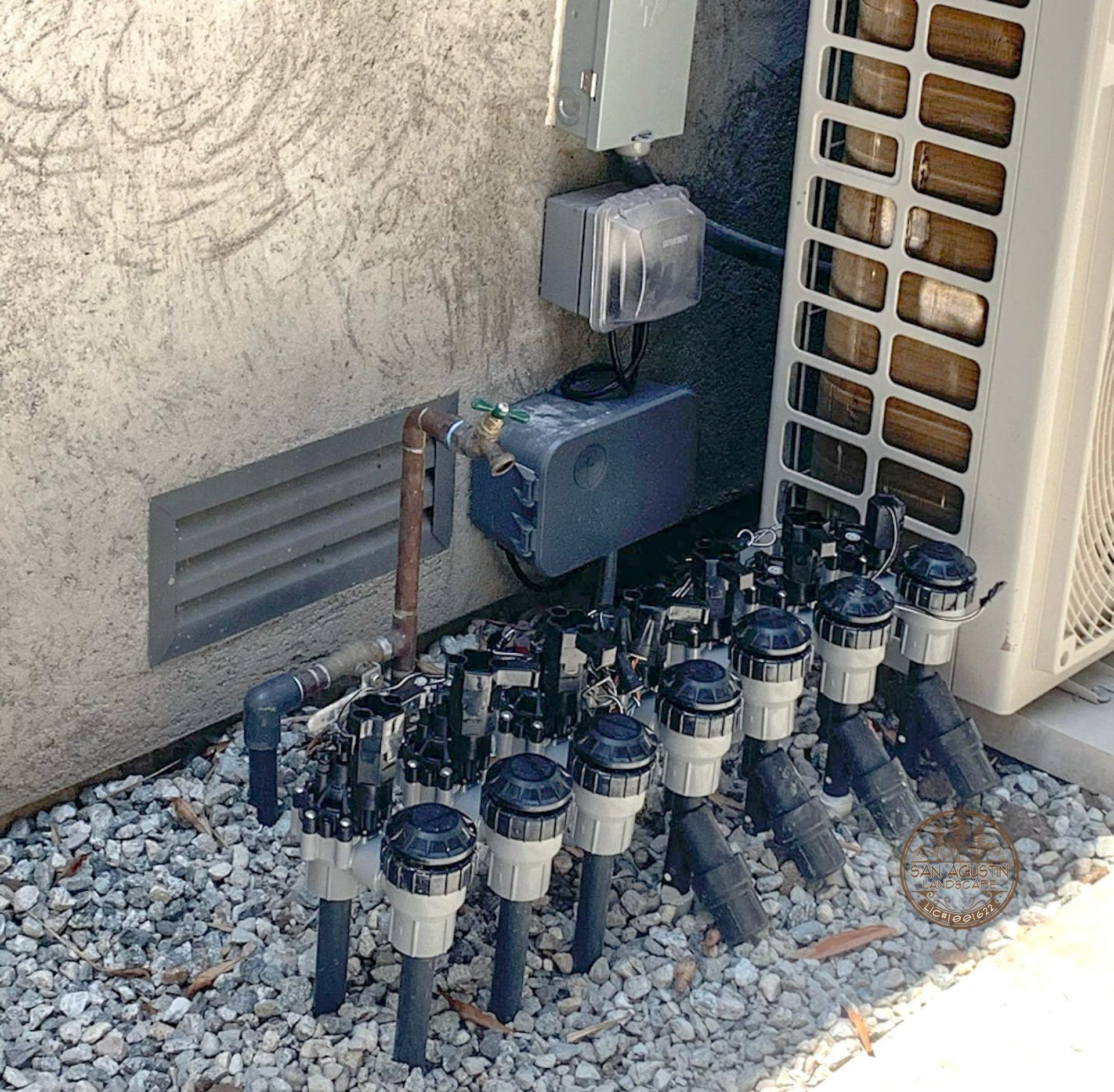 Plumbing system with multiple valves, electrical boxes, and an air conditioning unit against a stucco wall.