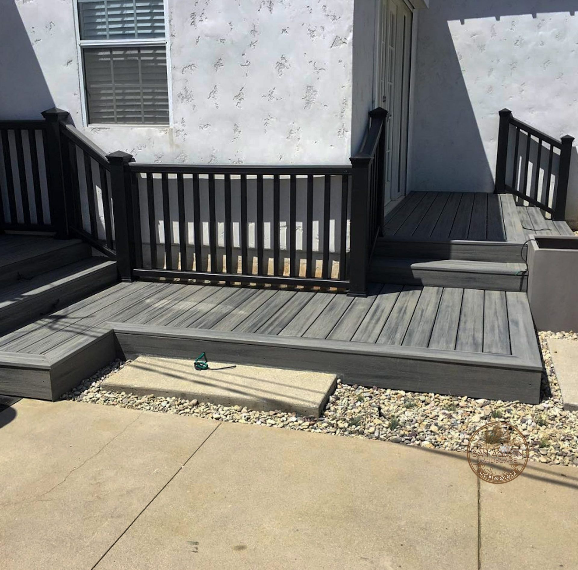 Gray wooden deck with steps, handrails, and a slatted fence, against a stucco wall.