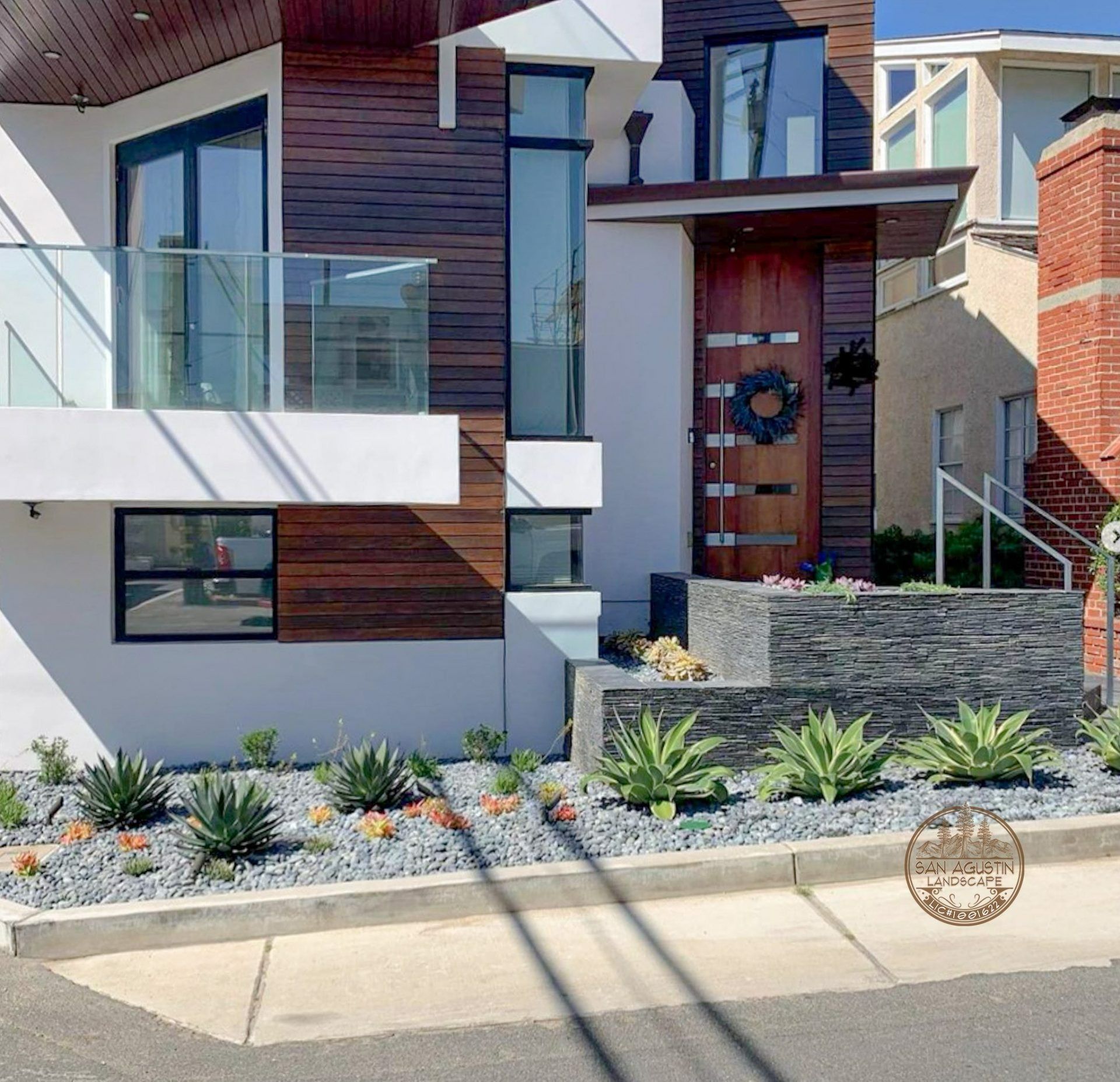 Modern home with wooden facade, glass balcony, and succulent landscaping along a concrete sidewalk.
