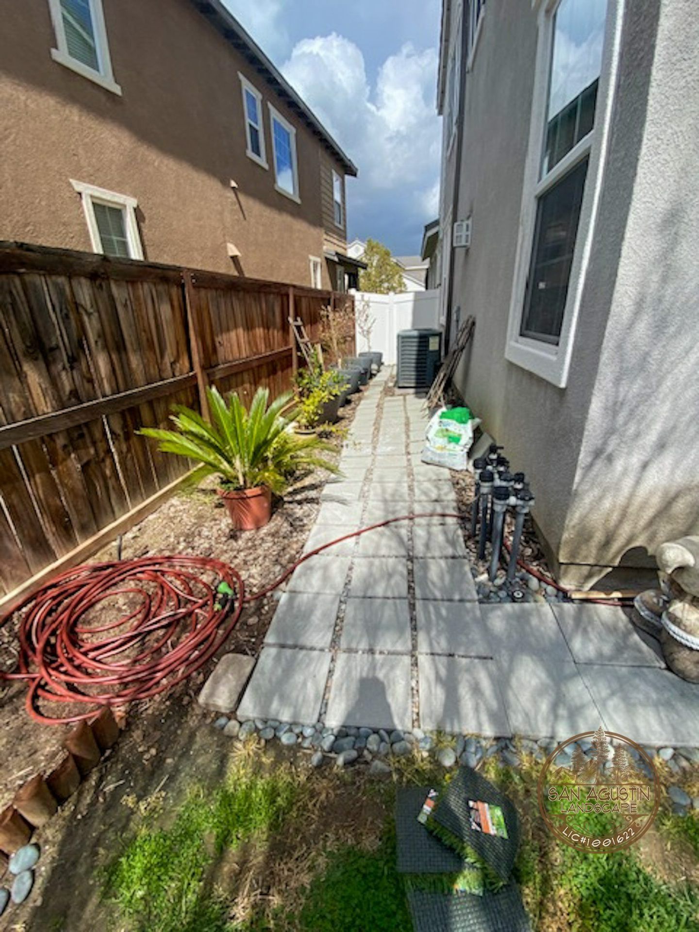 Narrow backyard path with pavers, surrounded by brown fence, beige house, plants, and hose.