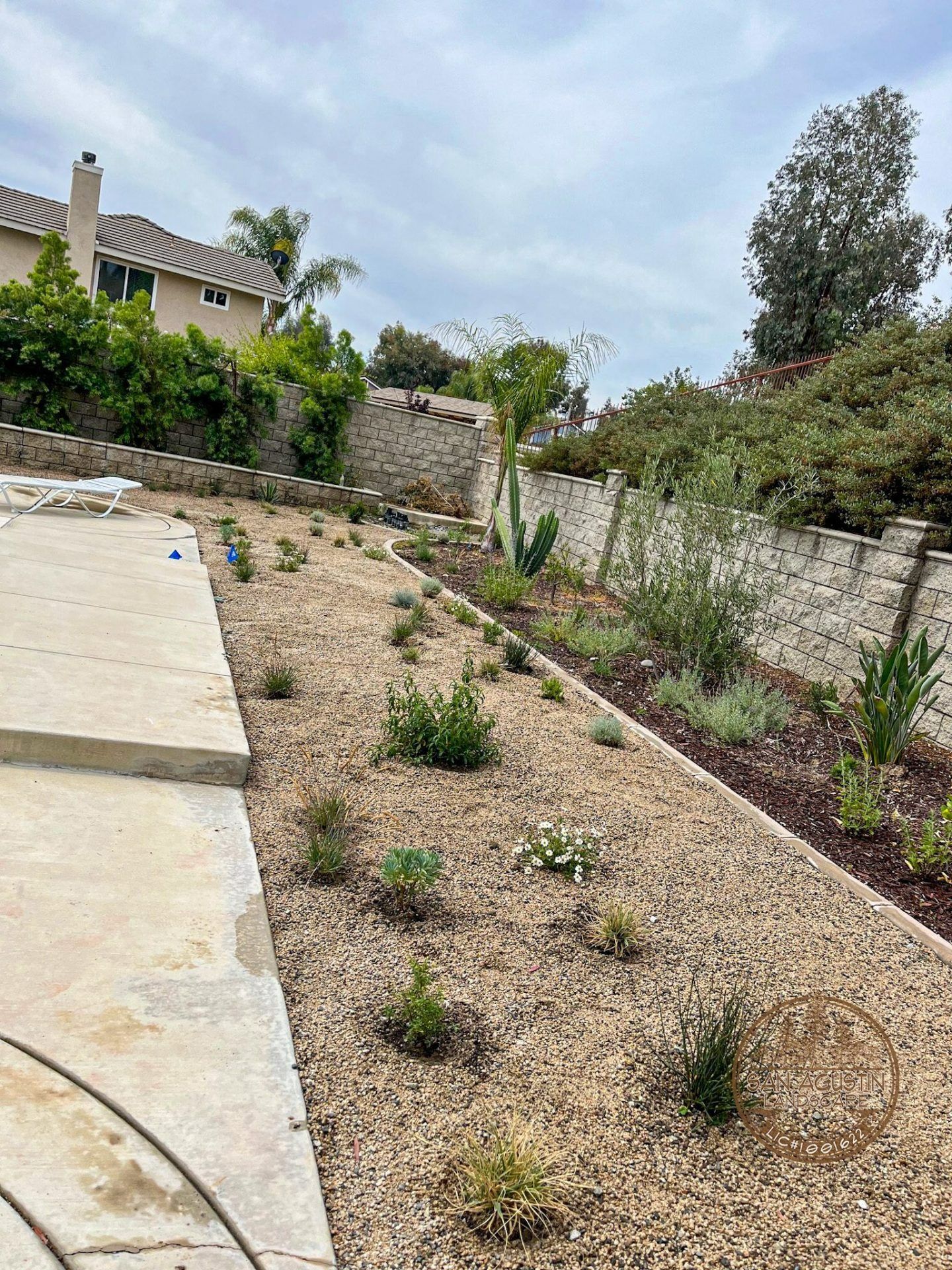 Sloped yard with gravel, young plants, stone retaining walls, and a house. Overcast sky.
