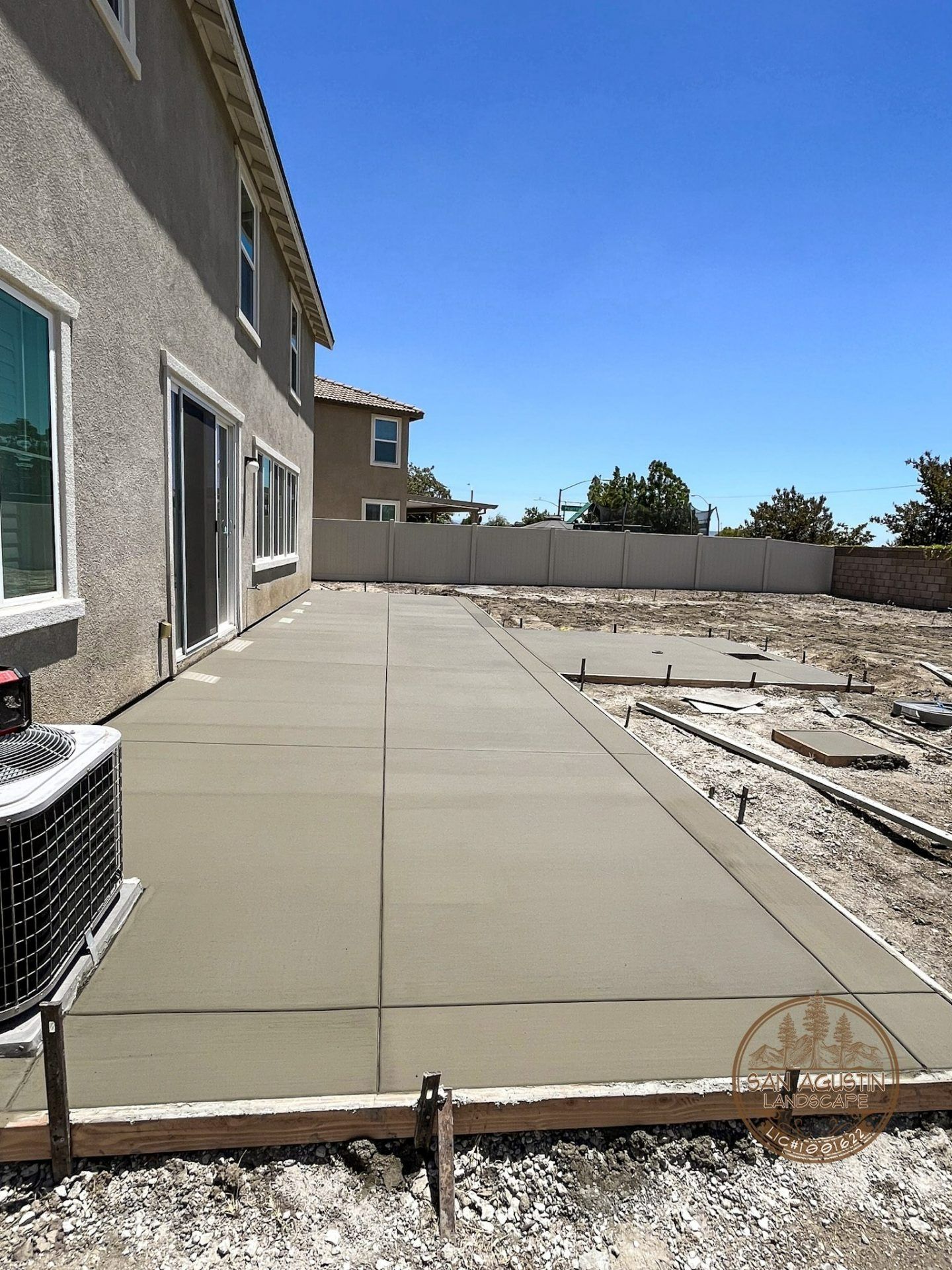 New concrete patio extension in a backyard, beside a two-story beige house under a blue sky.