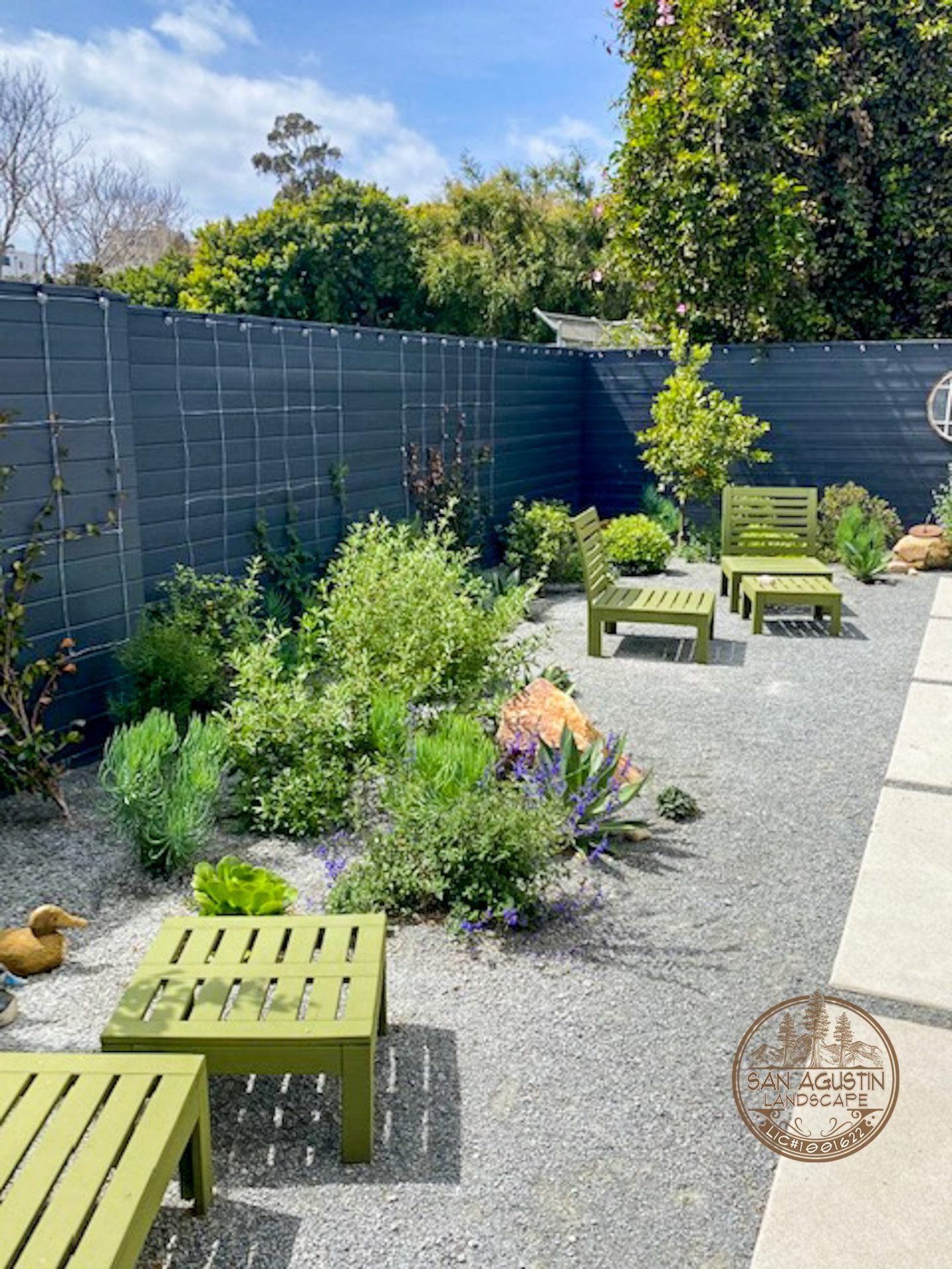 A gravel-covered backyard with green patio furniture. Plants and a dark gray fence are in the background.