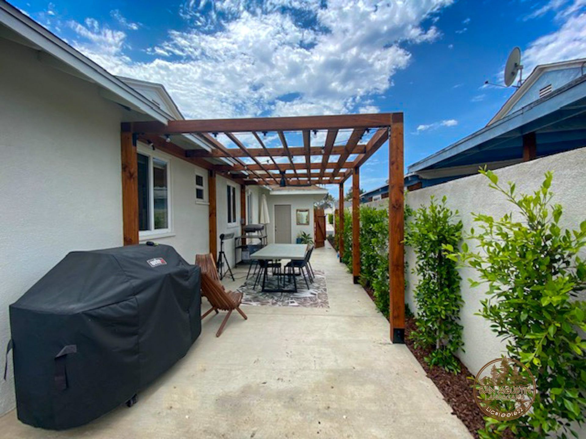 Outdoor patio with wooden pergola, dining set, and grill under a bright blue sky.