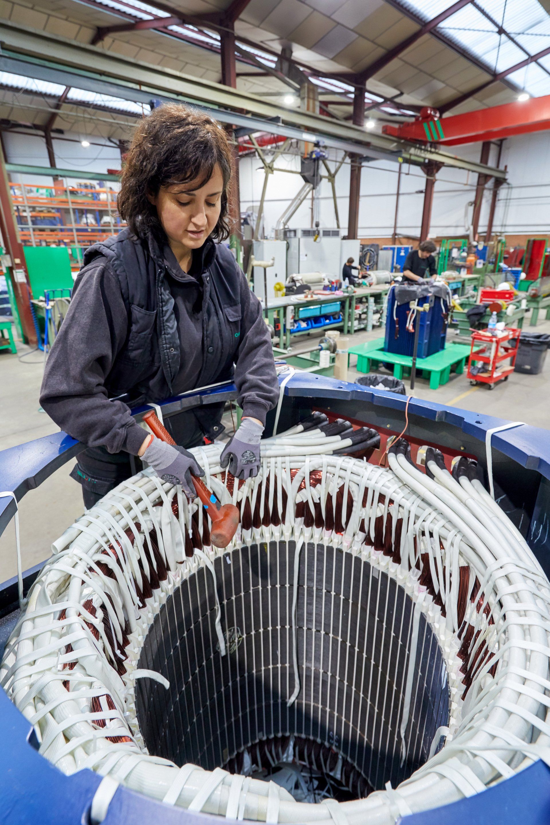 Mujer trabajando con un componente eléctrico grande en una fábrica, usando guantes y herramientas.