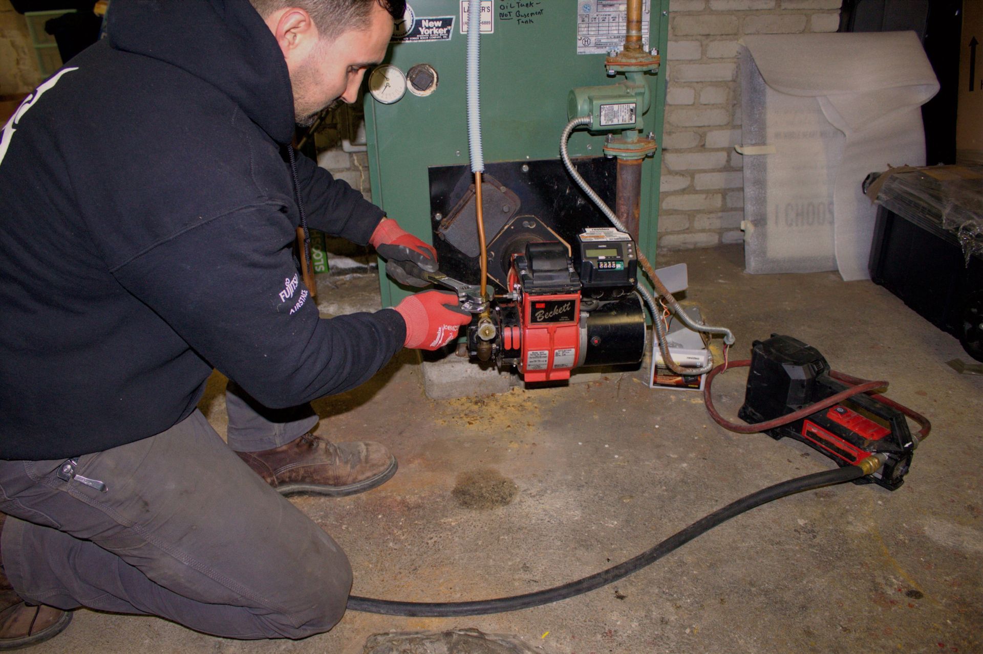 Technician kneeling beside a red boiler unit with piping and tools in a utility room