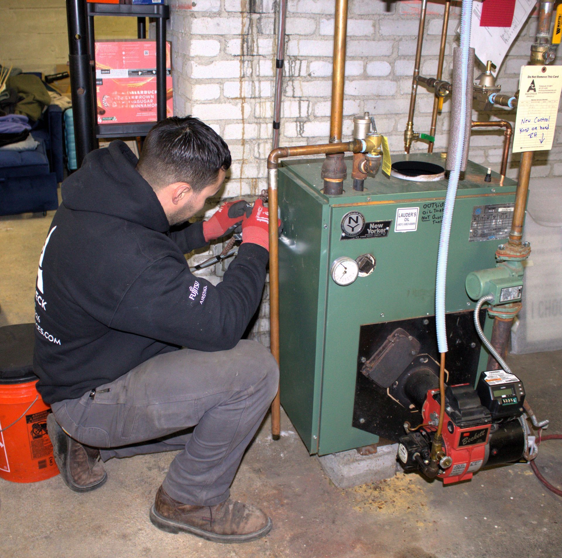 Technician kneels beside a green boiler, inspecting or repairing pipes in a basement.