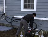 Person bending over a bicycle beside a gray house with cables along the wall