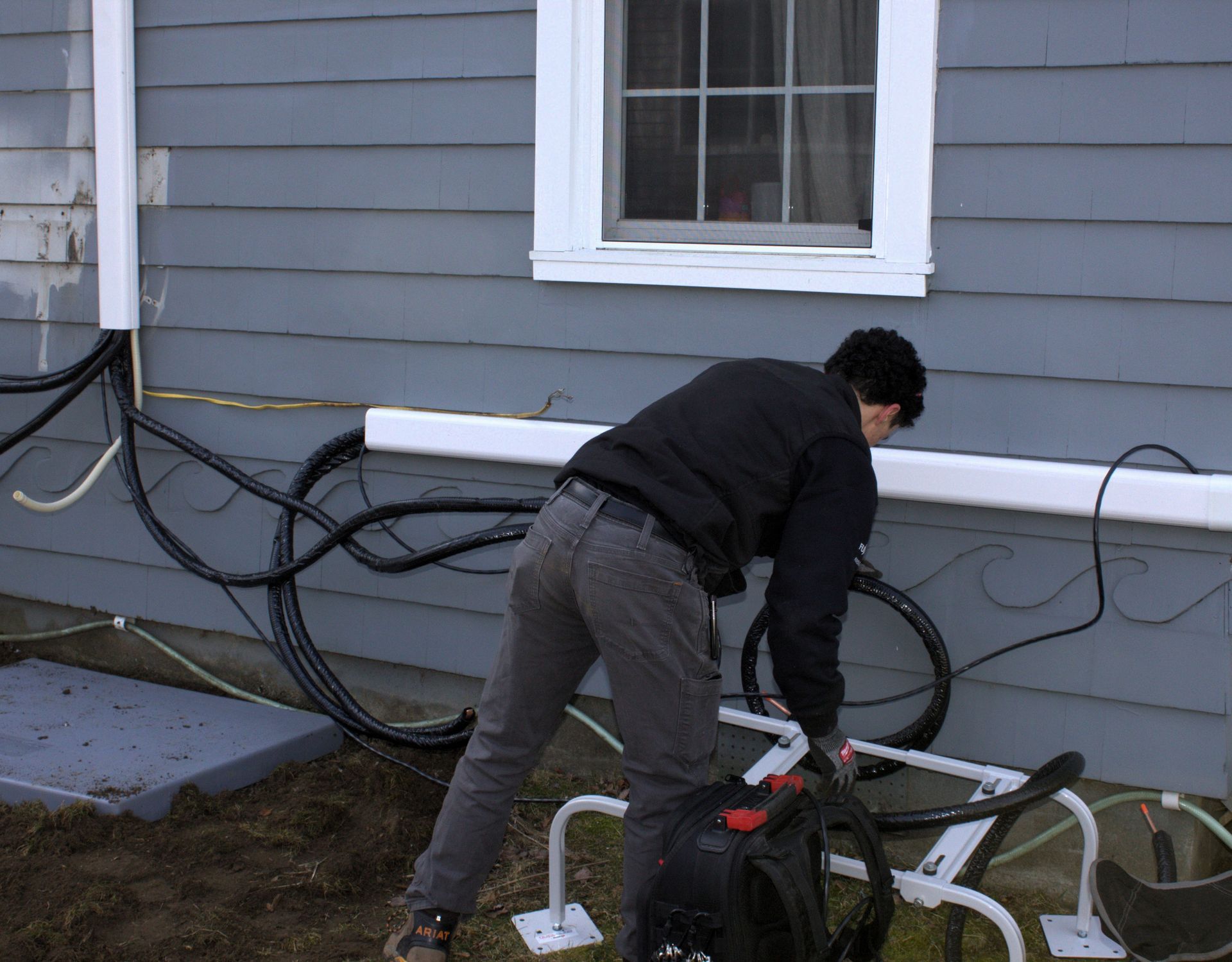 Person bending over a bicycle beside a gray house with cables along the wall