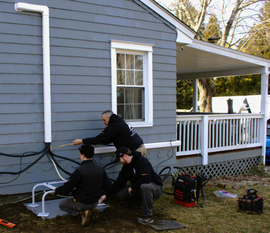 Three people kneel beside a house foundation, working on black cables and a white utility box.
