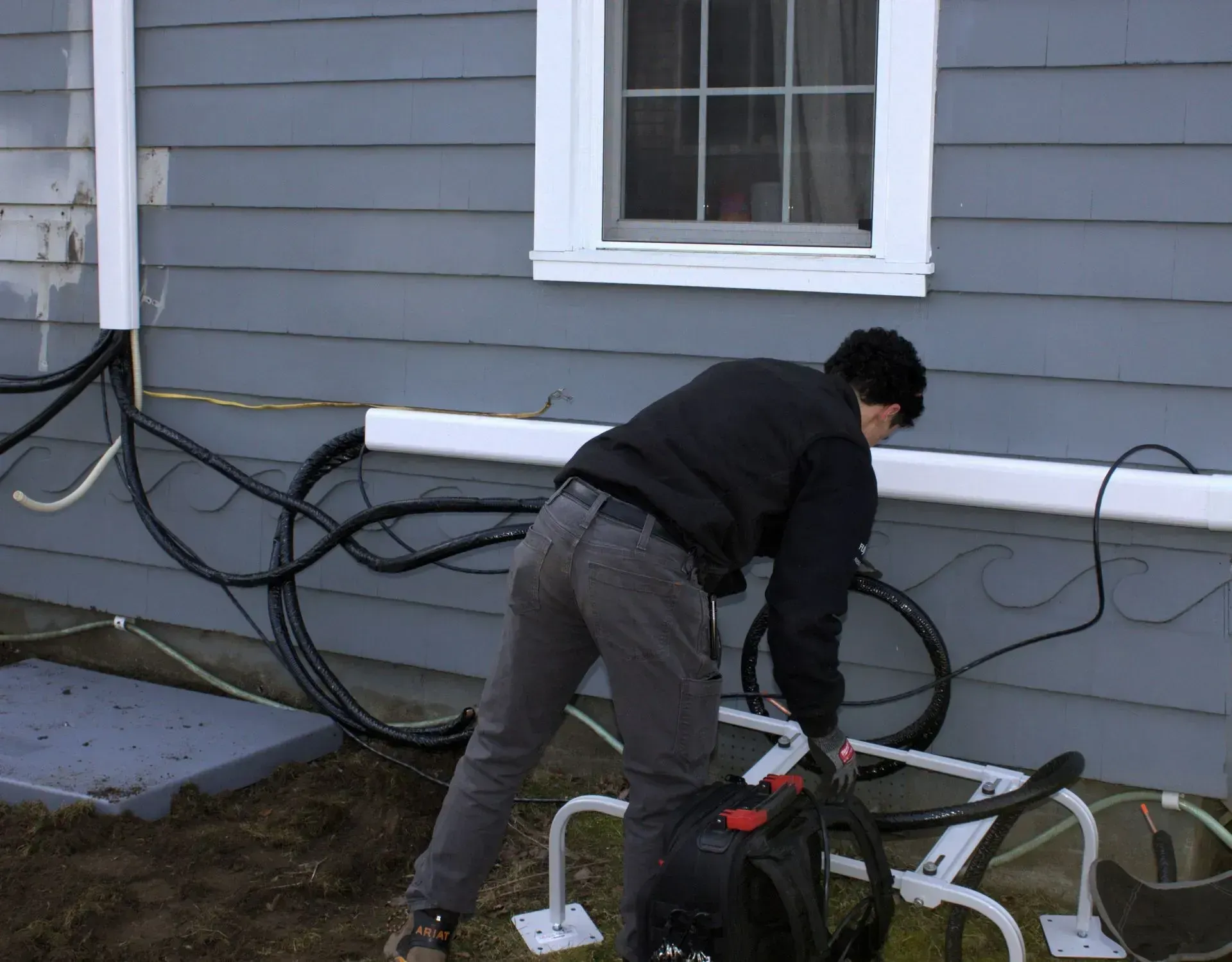 Person bending over a bicycle beside a gray house with cables along the wall