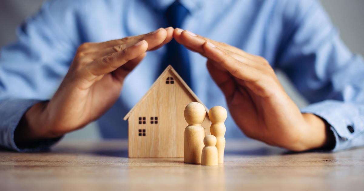 Hands shielding a small wooden house and family figurines on a table