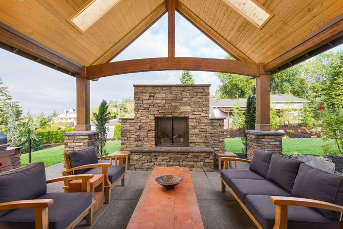 Covered outdoor patio with stone fireplace, wood beams, and cushioned seating around a fire pit.