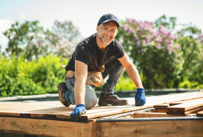 Smiling roofer kneels on a wooden roof frame outdoors, wearing a black cap, shirt, and blue gloves.