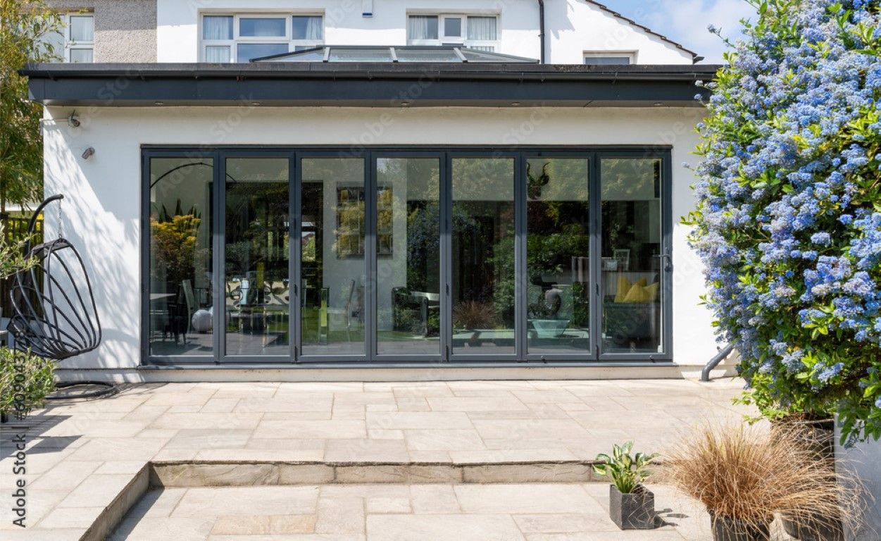 Modern glass-fronted house extension with wide sliding doors and a patio, framed by greenery and blue flowers