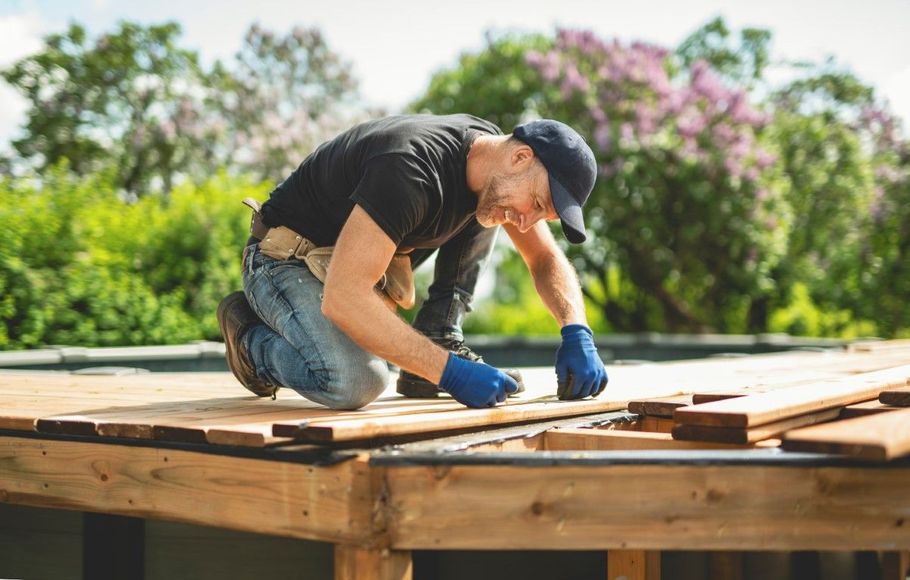 Worker in blue gloves kneeling on a wooden roof frame outdoors, measuring or fastening boards.