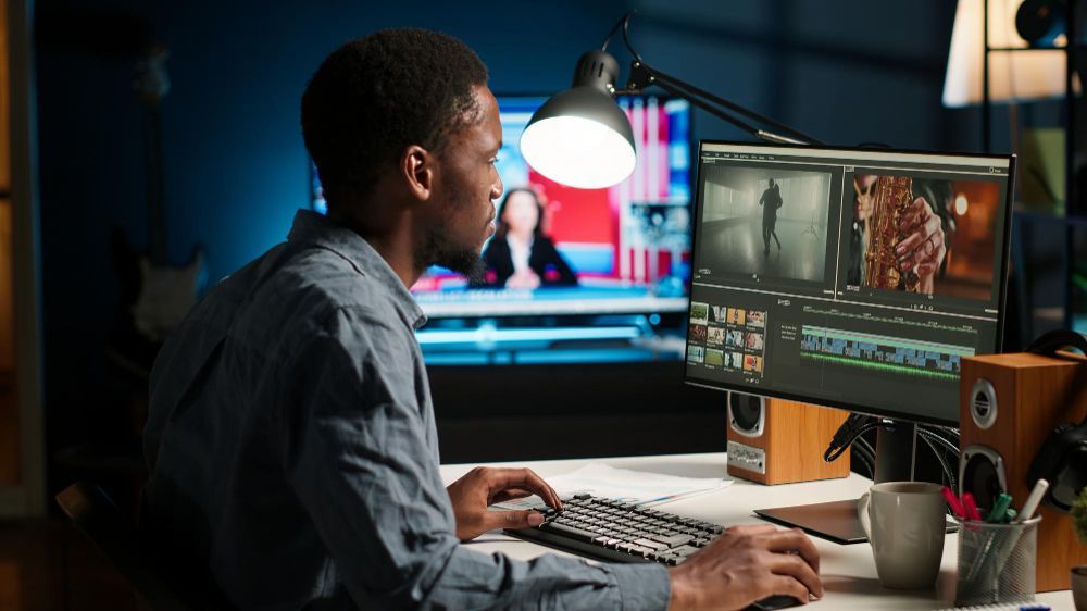 Person editing video on a desktop computer in a dimly lit workspace with dual monitors and a desk lamp