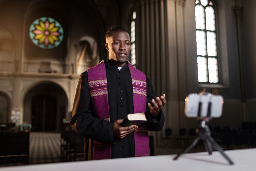 Priest in a purple stole speaking in a church, with a phone on a tripod in the foreground.