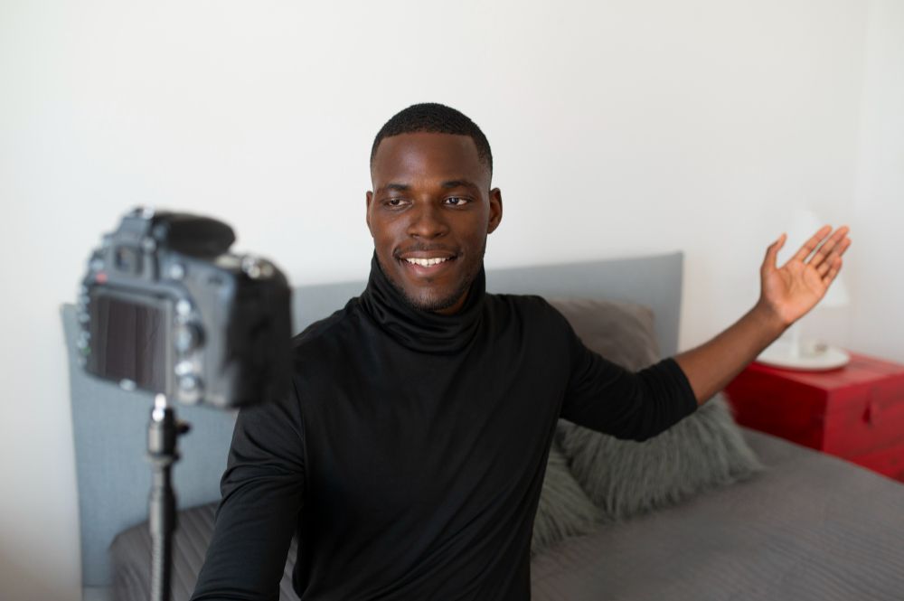 Man smiling and waving beside a camera on a tripod in a living room
