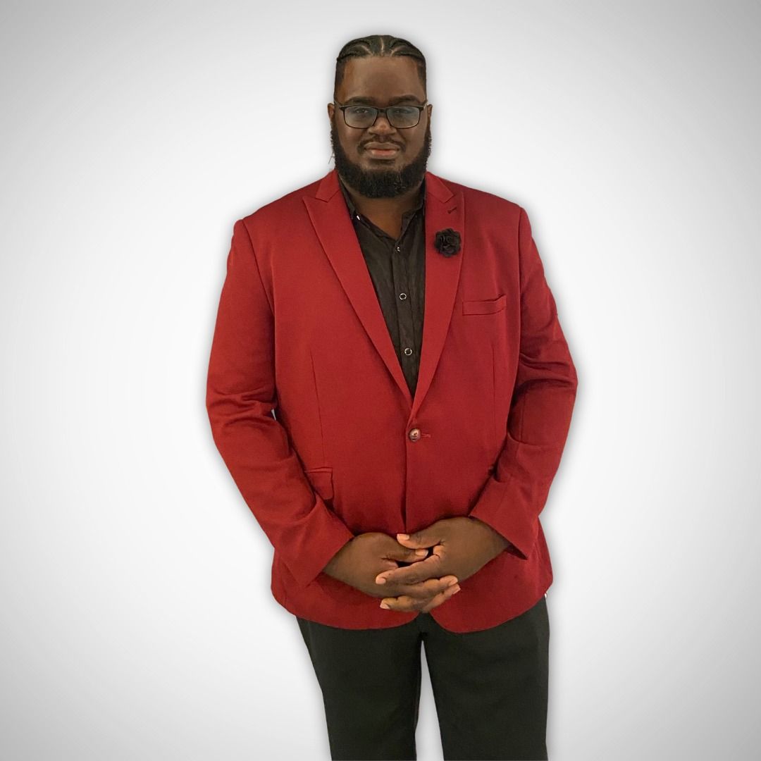 Man in a red blazer and black pants stands with hands clasped against a white background.