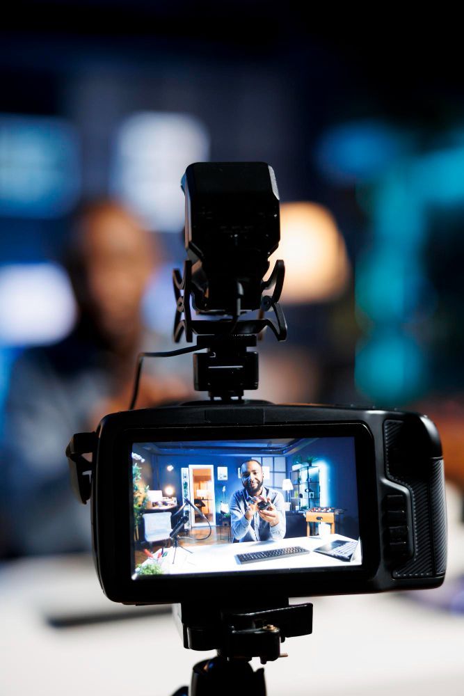 Video camera recording a presenter on a brightly lit studio set, with the camera screen in focus.