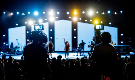 Concert stage with bright blue and yellow lights, performers onstage, and silhouetted audience in the foreground
