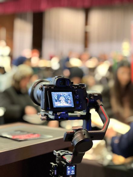 Camera on a tripod filming a blurred conference audience in a meeting room