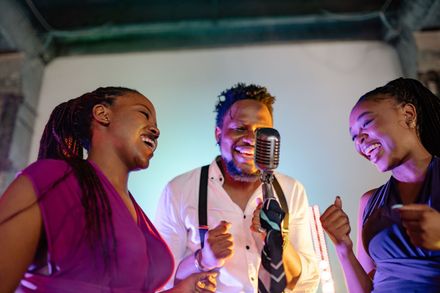 Three people singing into a vintage microphone on a stage, smiling under purple lighting