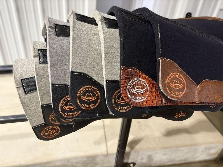 Horse saddle pads displayed on a metal rack. Various colors: gray, black, brown with logo.