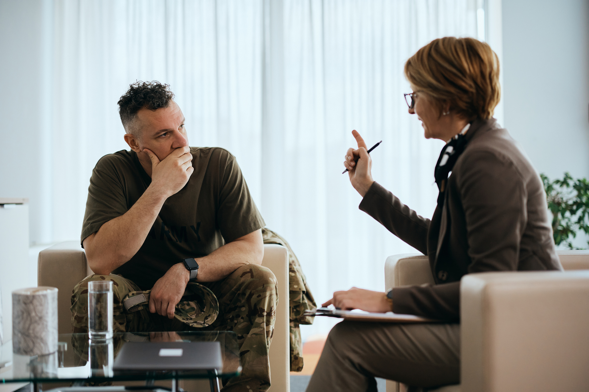Two people in a bright office meeting, one speaking and one listening across a table.