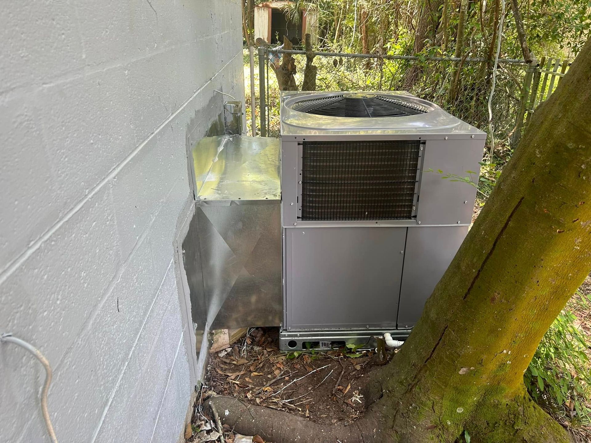 Outdoor HVAC unit beside a gray wall and tree, with metal ductwork and a coil on top